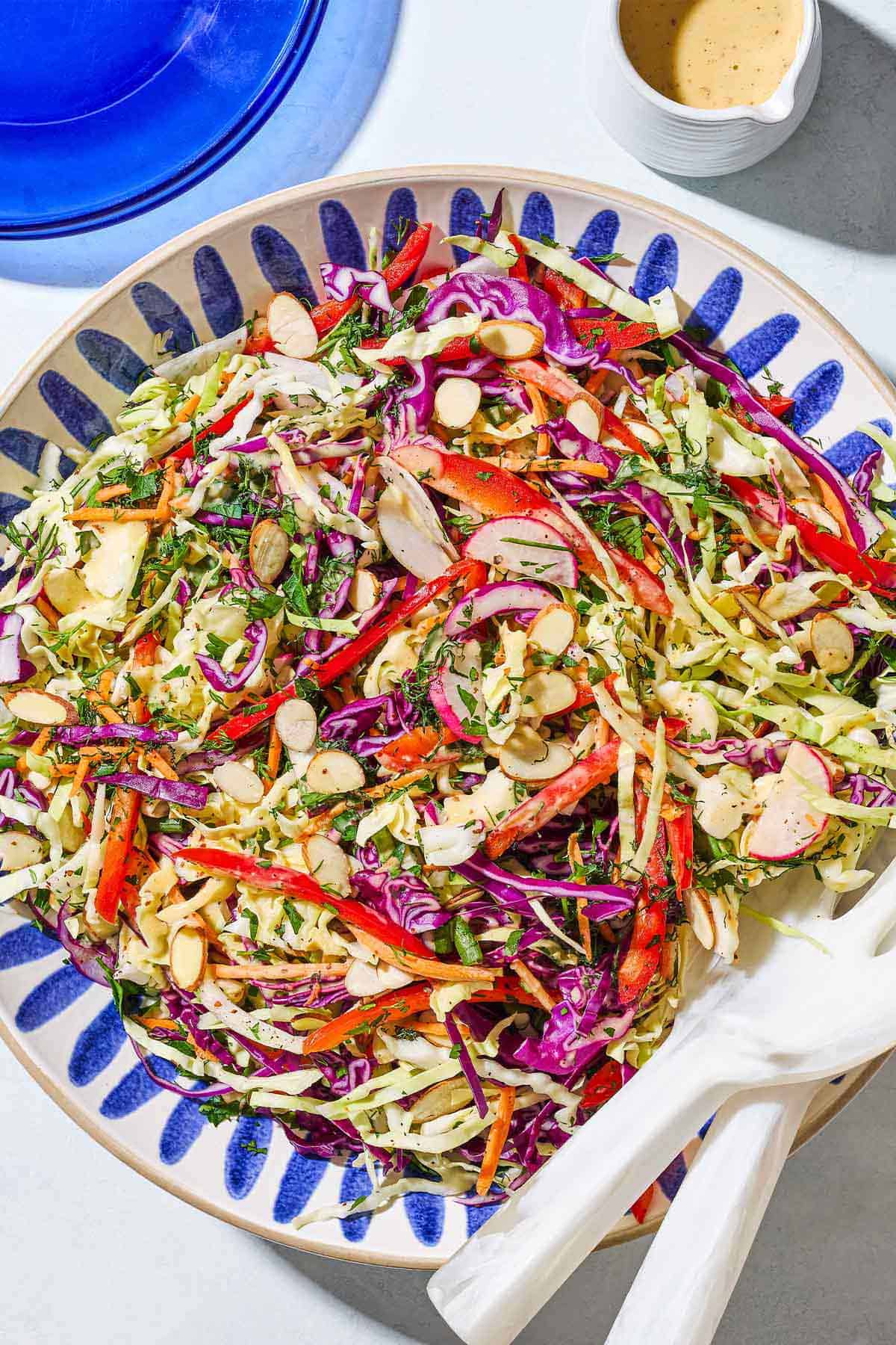 An overhead photo of cabbage salad in a serving bowl with serving utensils surrounded by a small container of the dressing and a blue plate.