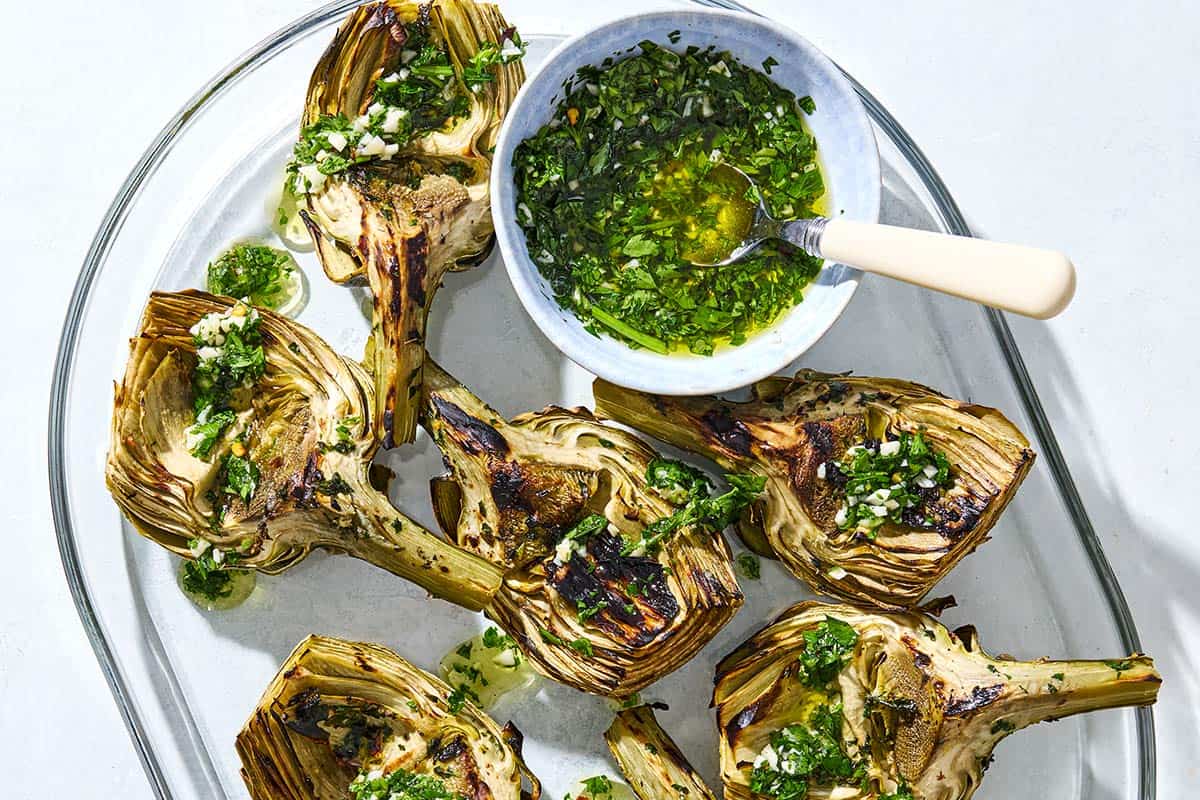 Grilled artichoke quarters drizzled with marinade on a serving platter along with some of the marinade in a bowl with a spoon.
