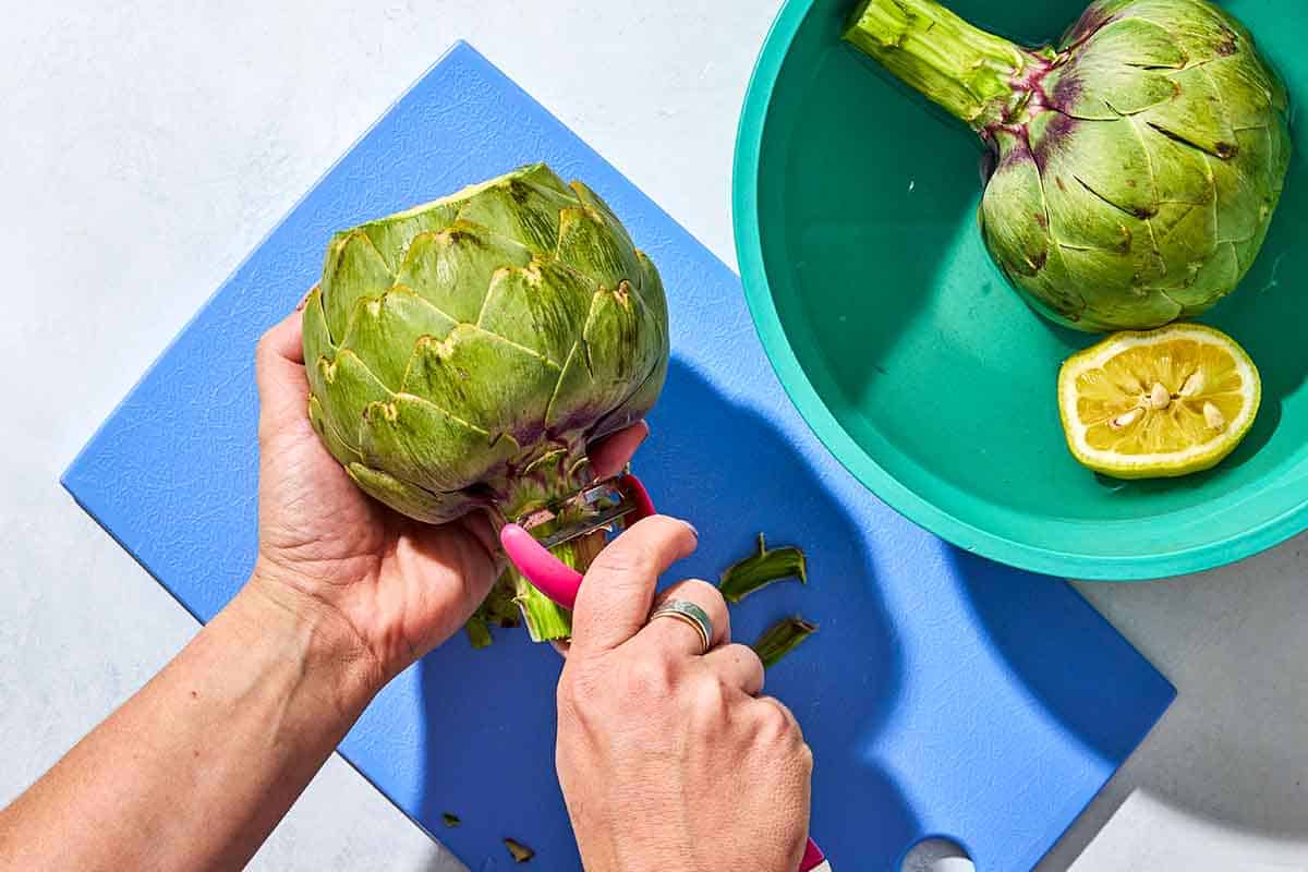 An artichoke being trimmed on a cutting board next to a bowl with another artichoke and a juiced lemon half.