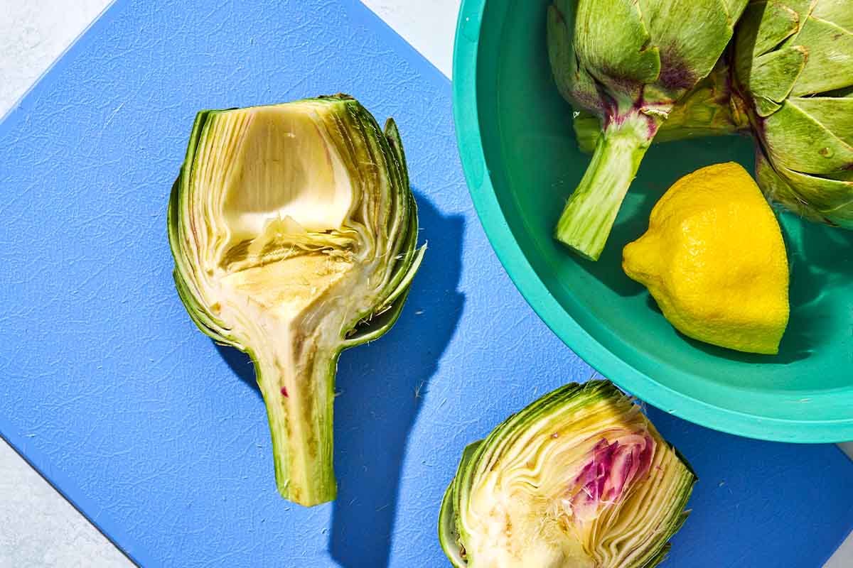 Two artichoke quarters being trimmed on cutting board next to a bowl with the rest of the artichoke quarters and a juiced lemon half.