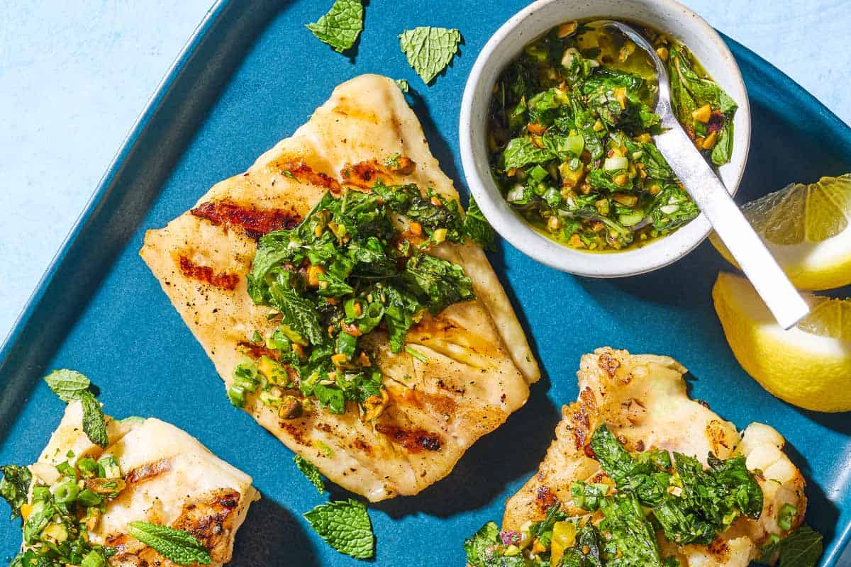 An overhead close up photo of 3 grilled cod filets topped with pistachio-herb salsa on a serving platter along with a small bowl of the salsa and 2 lemon wedges.