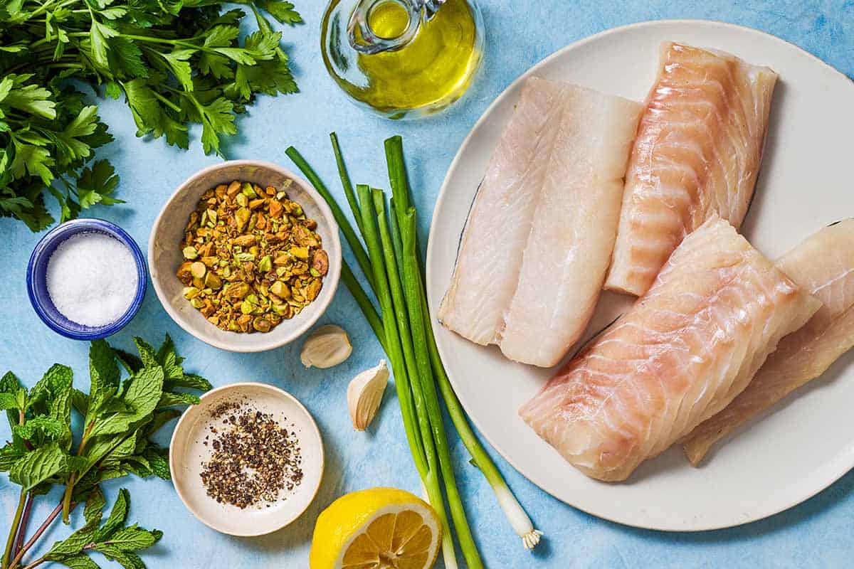 Ingredients for grilled cod including skin-on cod filets, shelled pistachios, parsley, mint leaves, green onions, garlic, lemon, olive oil salt and black pepper.