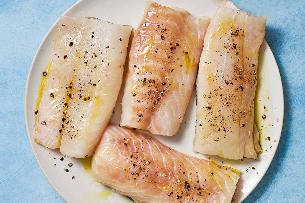 4 uncooked cod filets on a plate, brushed with olive oil and seasoned with salt and black pepper.