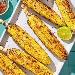 An overhead photo of 6 grilled corncobs on a serving platter with 2 lime halves. Next to this is a small bowl of Aleppo pepper.