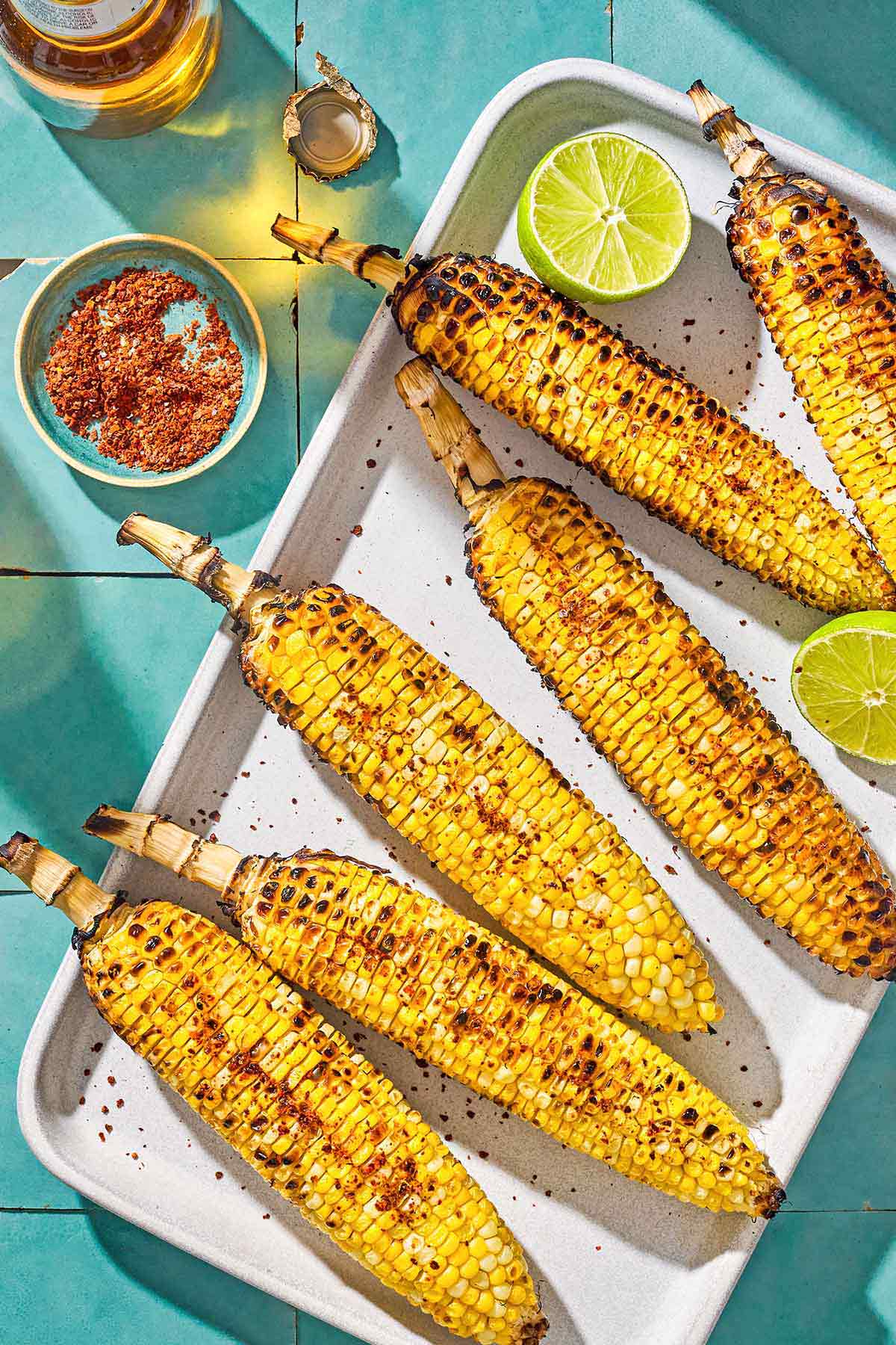 An overhead photo of 6 grilled corncobs on a serving platter with 2 lime halves. Next to this is a small bowl of Aleppo pepper and a bottle of beer.