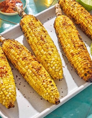 An overhead photo of 5 grilled corncobs on a serving platter with lime half. Next to this is a small bowl of Aleppo pepper.