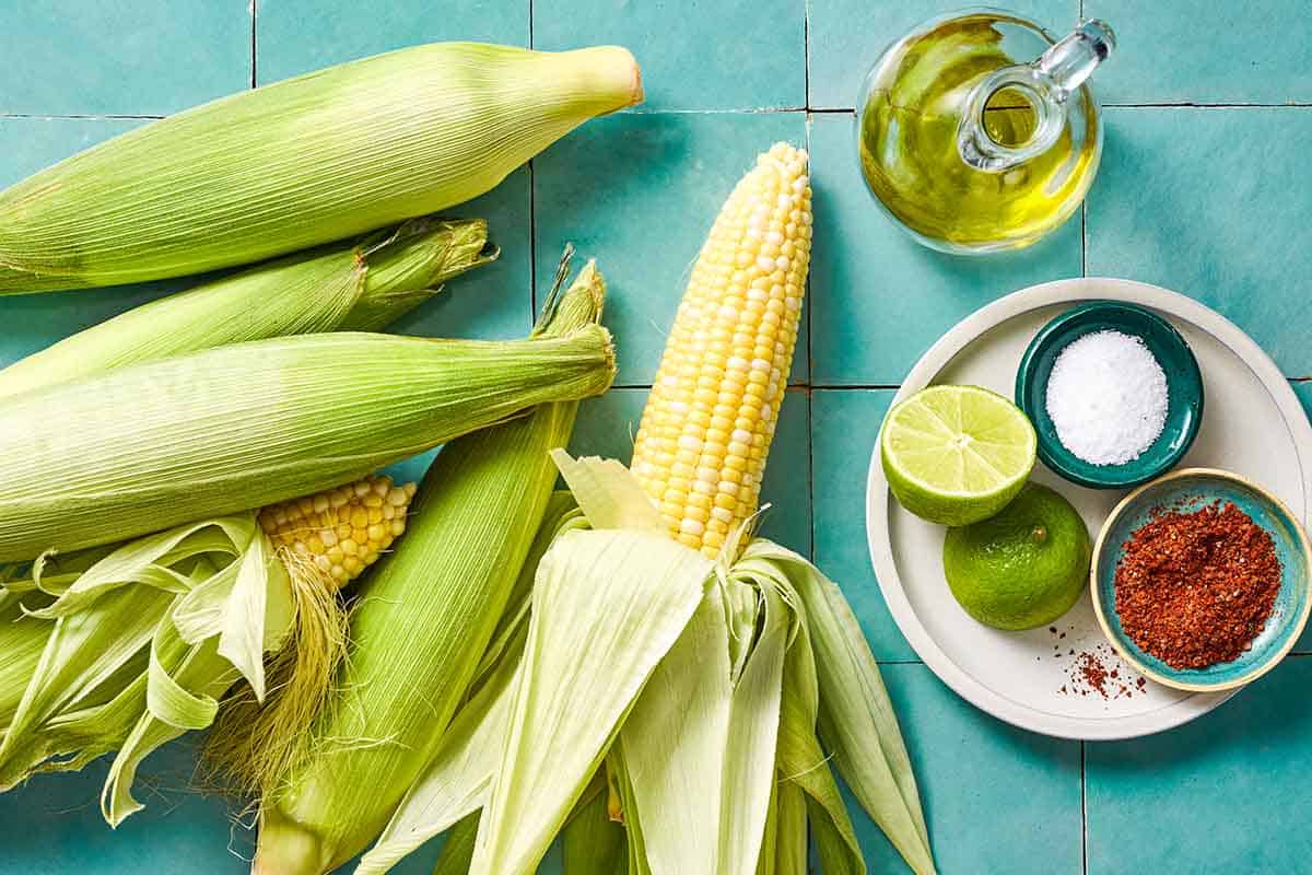 Ingredients for grilled corn on the cob including 5 ears of corn, a bottle of olive oil, and a plate containing 2 lemon halves and bowls of kosher salt and Aleppo pepper.