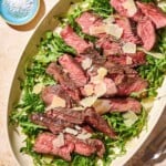 An overhead photo of beef tagliata topped with shaved parmesan on a bed of arugula on a serving platter. Next to this is a small bowl of salt and a serving fork.