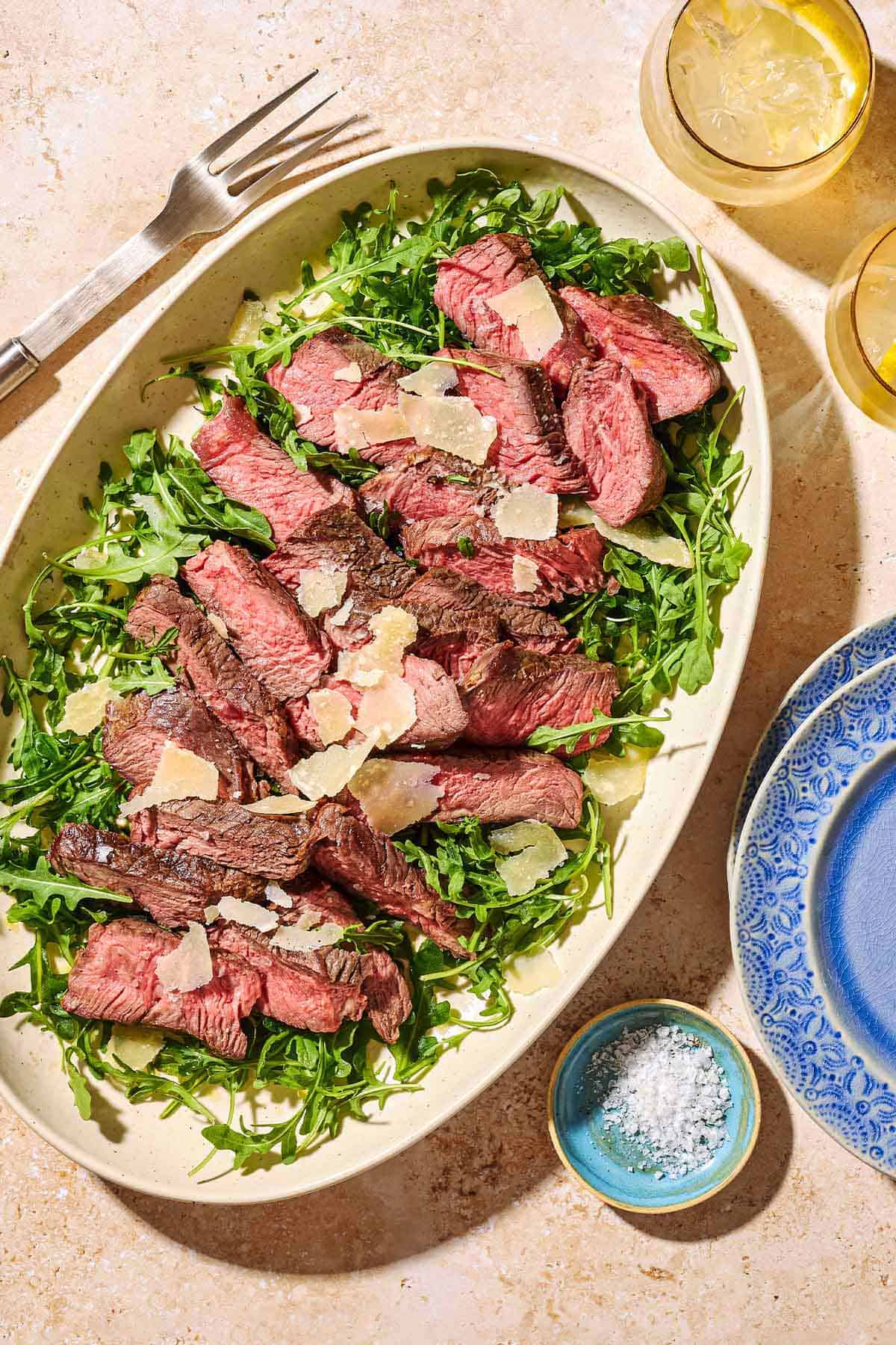 An overhead photo of beef tagliata topped with shaved parmesan on a bed of arugula on a serving platter. Next to this is a small bowl of salt, two glasses of water, a stack of 2 plates, and a serving fork.