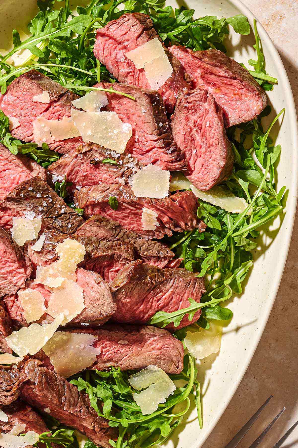 A close up of beef tagliata topped with shaved parmesan on a bed of arugula on a serving platter.