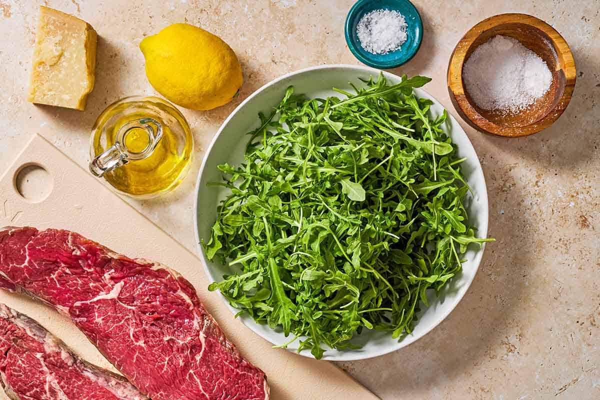 Ingredients for beef tagliata including sirloin steak, olive oil, sea salt, flaky salt, arugula, lemon, and parmesan cheese.