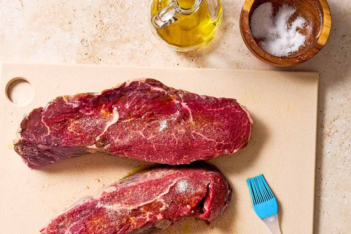 Two uncooked sirloin steaks brushed with olive oil and sprinkled with salt. Next to this is a bottle of olive oil and a bowl of salt.