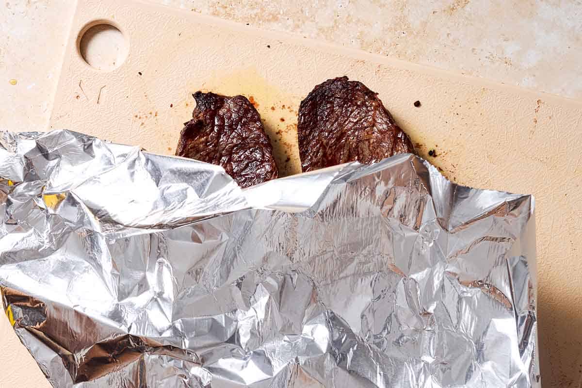 Two grilled sirloin steaks on a cutting board, partially covered with foil.