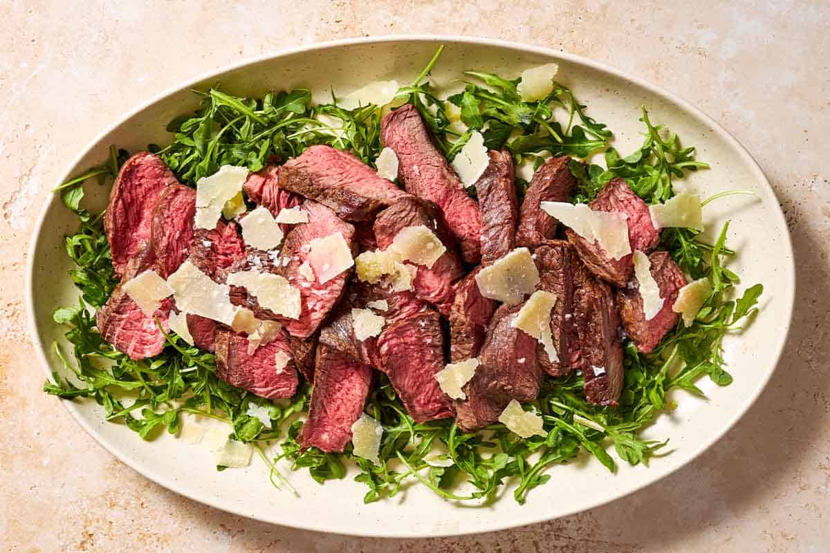 An overhead photo of beef tagliata topped with shaved parmesan on a bed of arugula on a serving platter.
