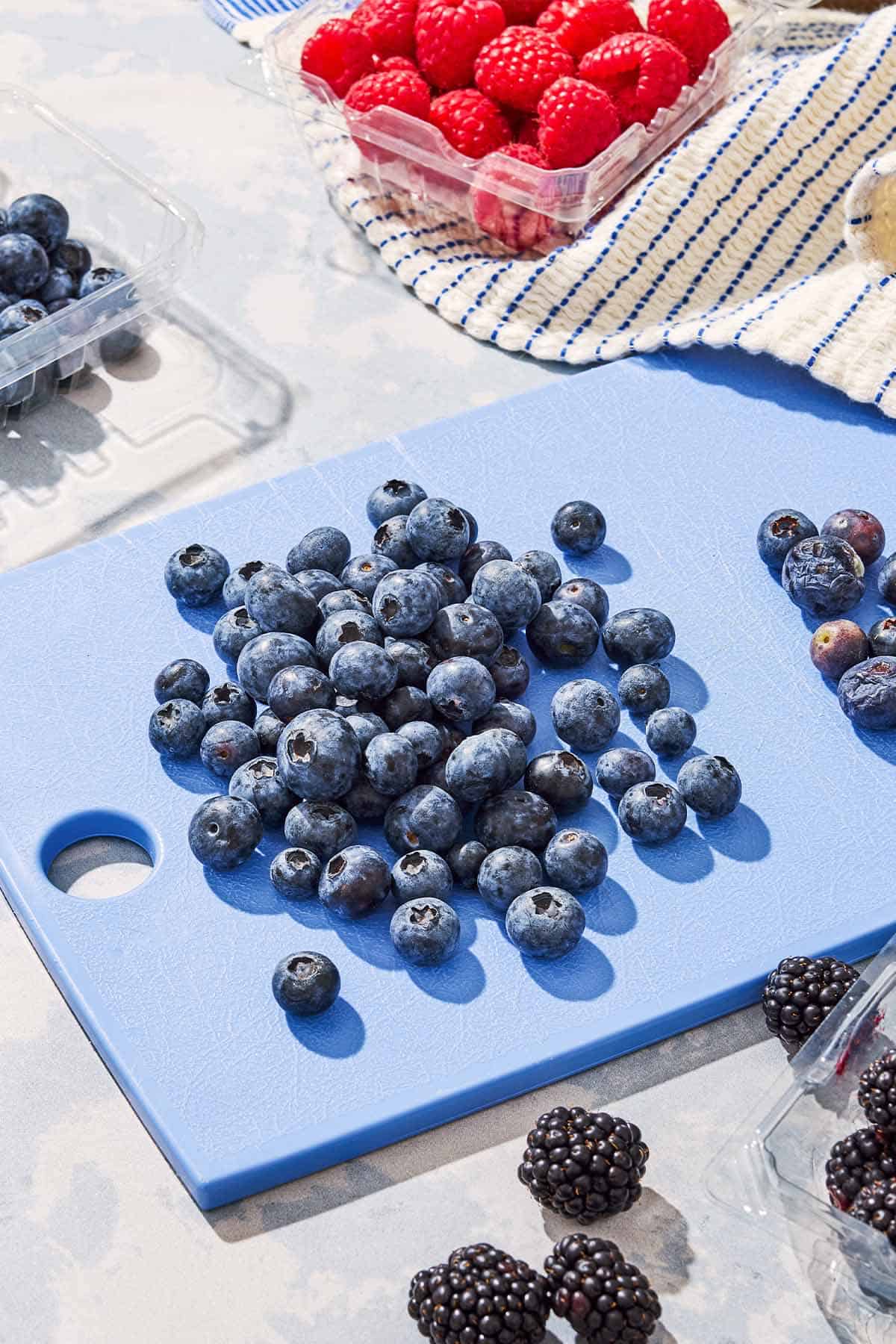 A handful of blueberries on a cutting board. This is surrounded by packs of raspberries, blackberries and more blueberries.