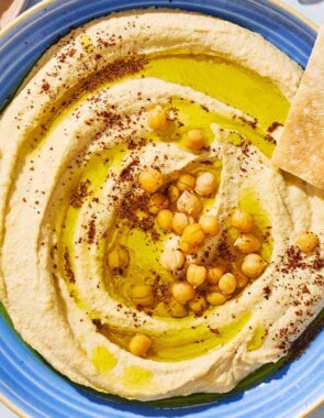 An overhead photo of hummus in a bowl topped with olive oil, chickpeas and sumac. Surrounding this is a bowl of sliced cucumber and pieces of pita bread.