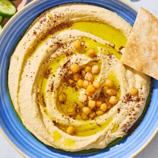 An overhead photo of hummus in a bowl topped with olive oil, chickpeas and sumac. Surrounding this is a bowl of sliced cucumber and pieces of pita bread.