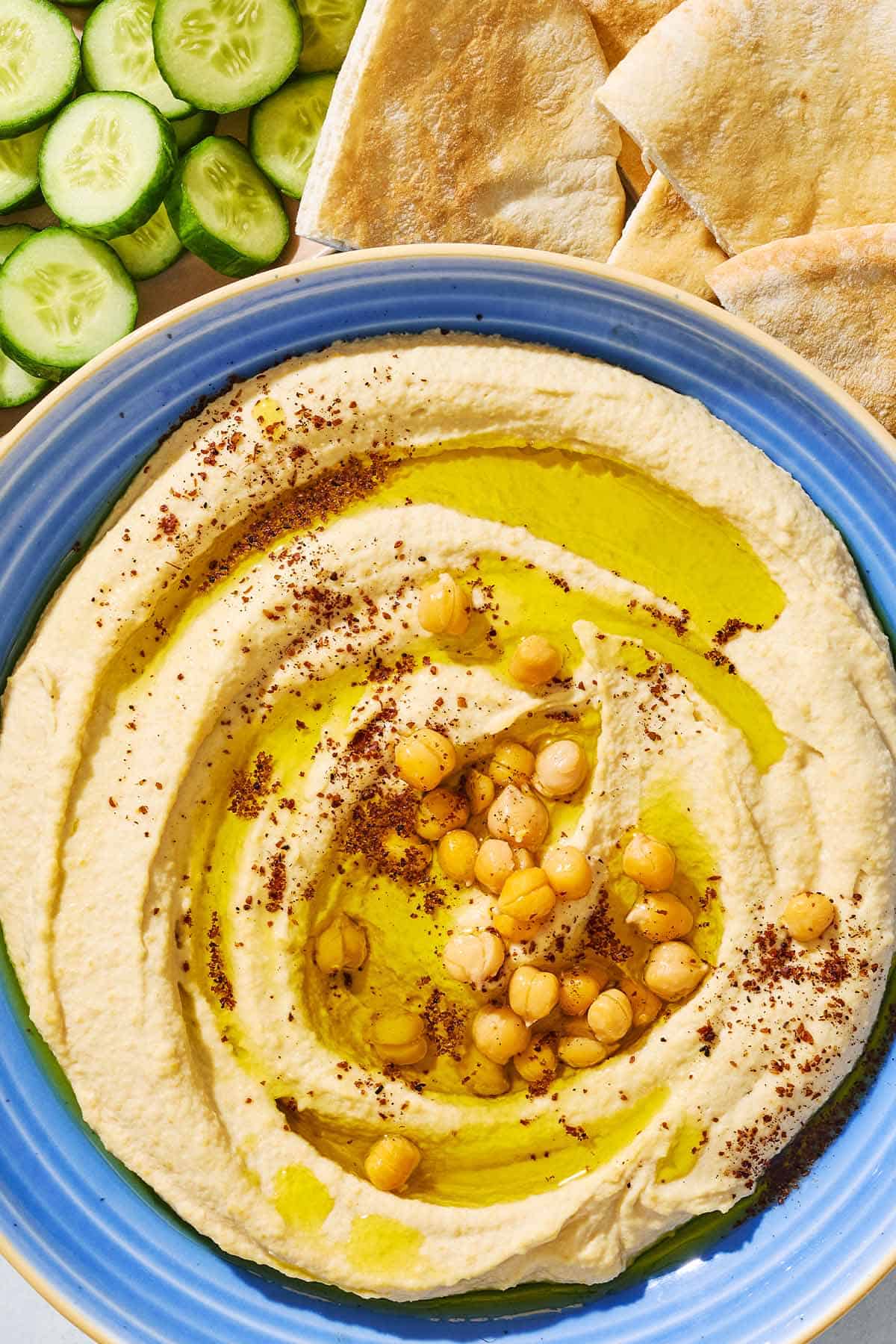 A close up of hummus in a bowl topped with olive oil, chickpeas and sumac. In the background is a bowl of sliced cucumber and pieces of pita bread.