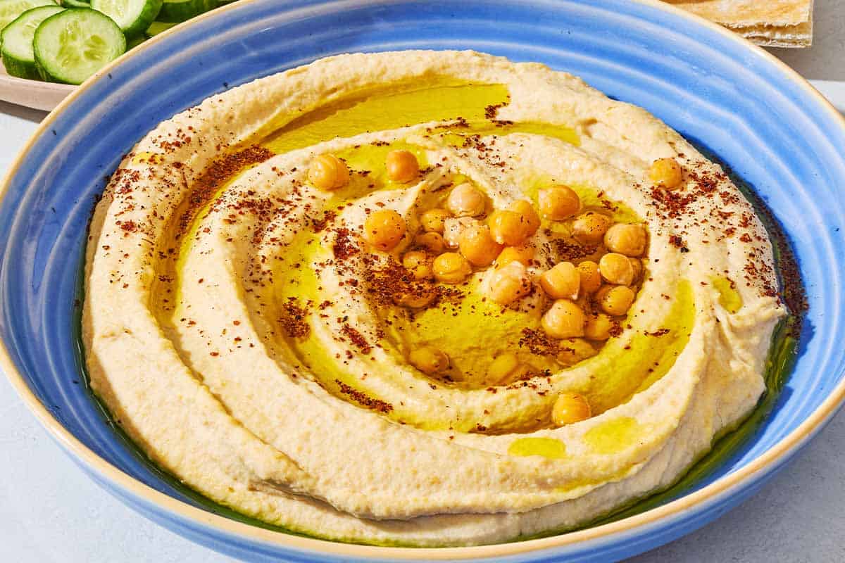 A close up of hummus in a bowl topped with olive oil, chickpeas and sumac. In the background is a bowl of sliced cucumber and pieces of pita bread.