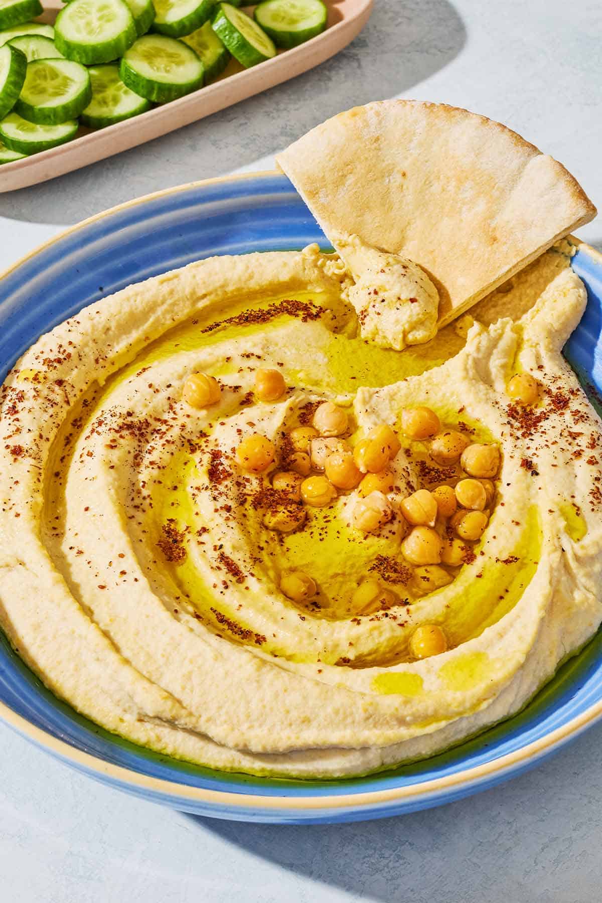 A close up of hummus in a bowl topped with olive oil, chickpeas and sumac along with a wedge of pita bread. In the background is a bowl of sliced cucumber.