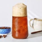 A close up of Freddo Espresso in a glass next to a bowl of coffee beans, a small pitcher and a spoon.