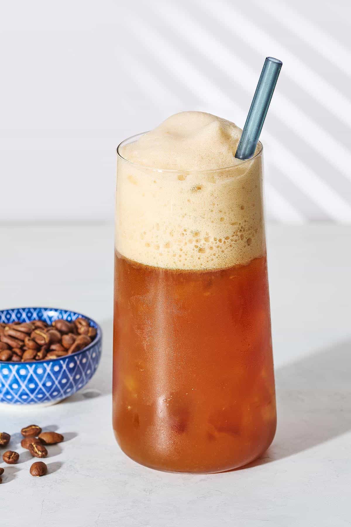 A close up of Freddo Espresso in a glass with a straw next to a bowl of coffee beans and some coffee beans on the table.