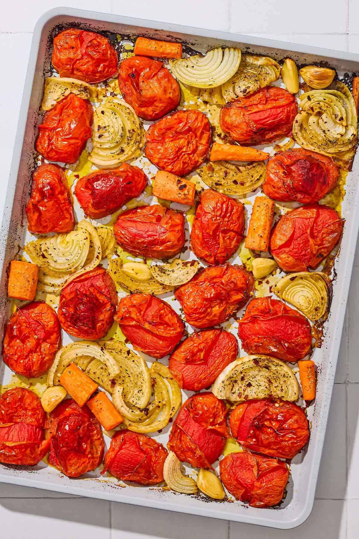 An overhead photo of roasted onions, tomatoes, carrots and garlic on a baking sheet.