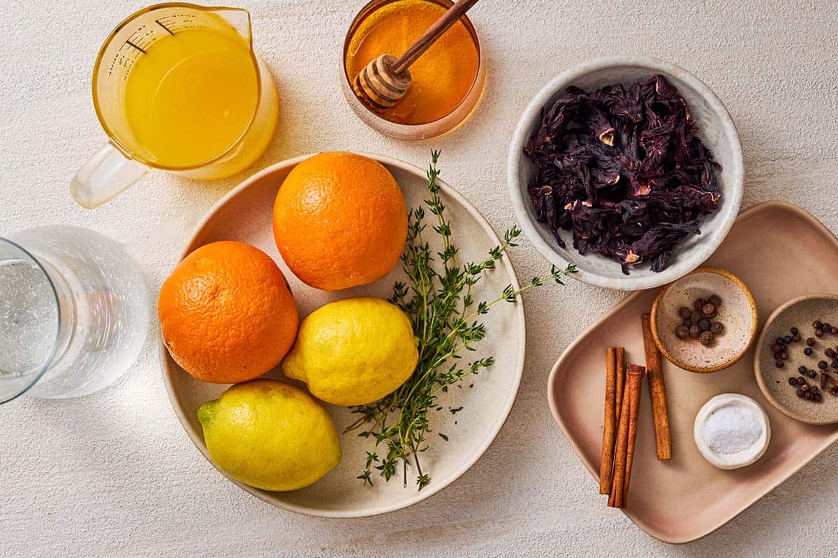 Ingredients for non alcoholic sangria including oranges, lemons, water, cinnamon sticks, fresh thyme, dried hibiscus flowers, honey, whole allspice, cloves, whole black peppercorns, and salt.