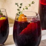 A close up of a glass of non alcoholic sangria garnished with sprigs of fresh thyme and orange and lemon peels on a marble serving tray.