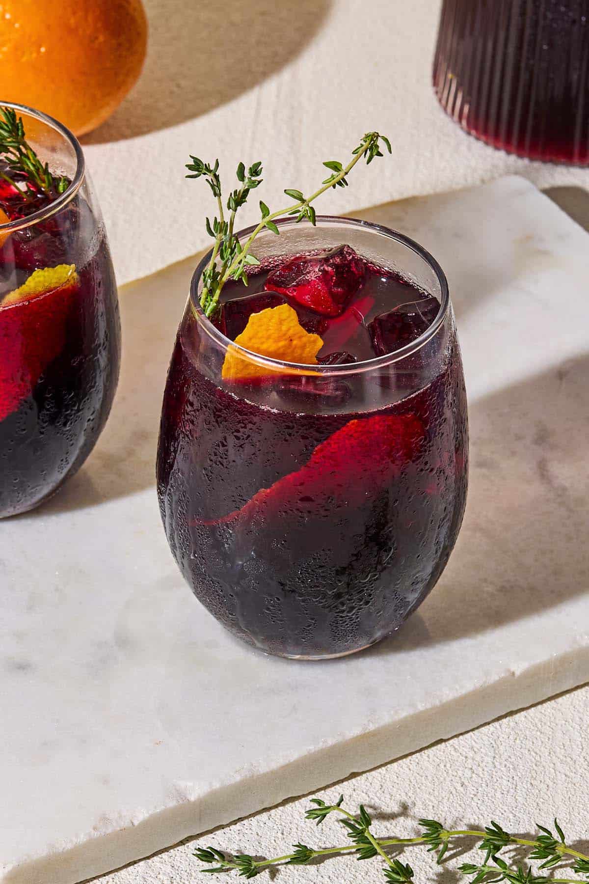 A close up of a glass of non alcoholic sangria garnished with sprigs of fresh thyme and orange and lemon peels on a marble serving tray.