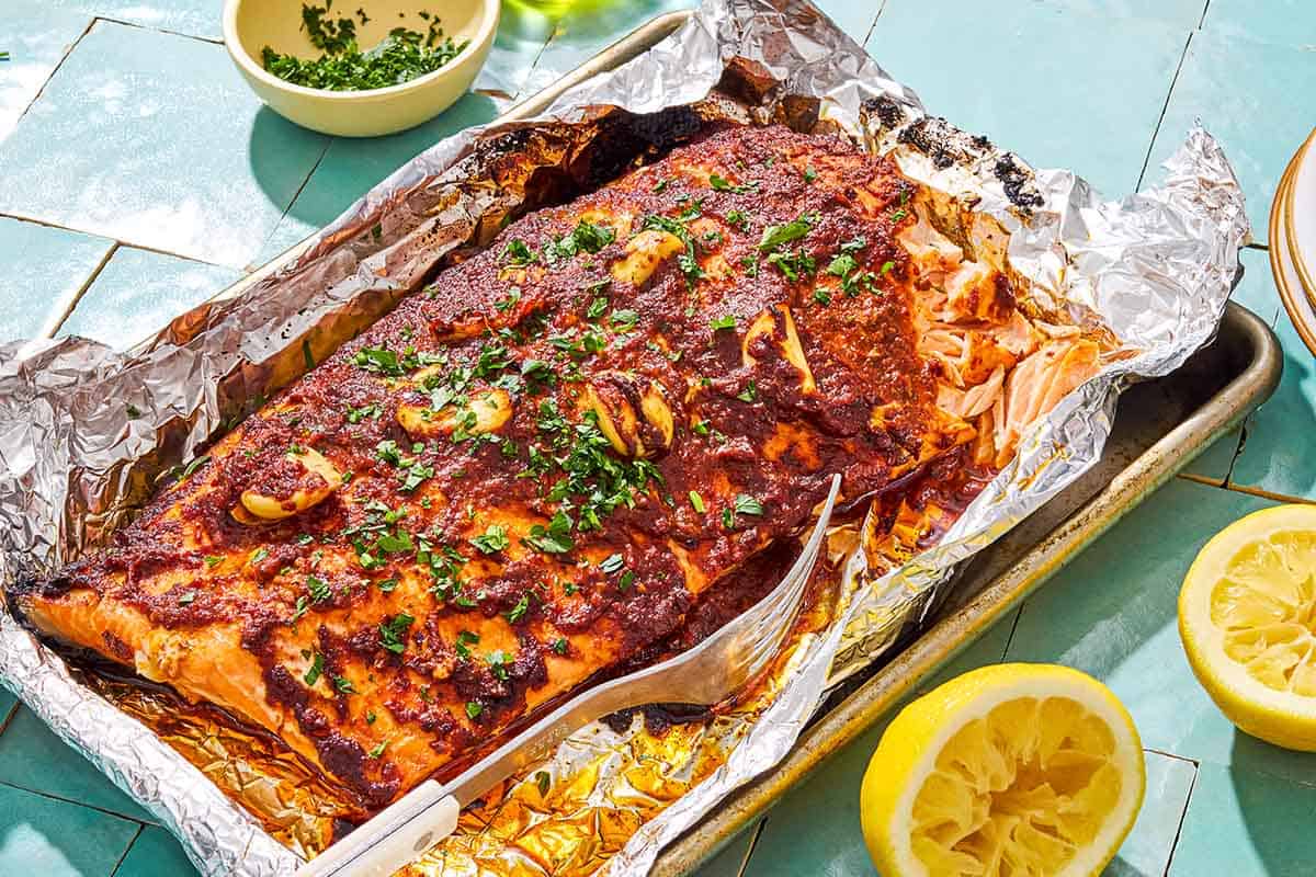 Baked Turkish style salmon on a foil-lined baking sheet with a fork. Next to this is a bowl of chopped fresh herbs, a stack of 2 plates, and 2 juiced lemon halves.