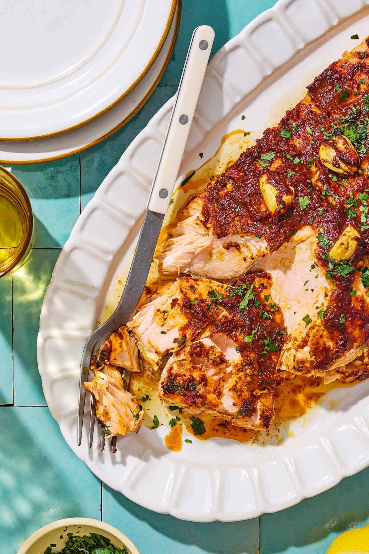 An overhead photo of Turkish style salmon on a serving platter with a fork surrounded by a stack of 2 plates, a glass of water and a small bowl of chopped fresh herbs.