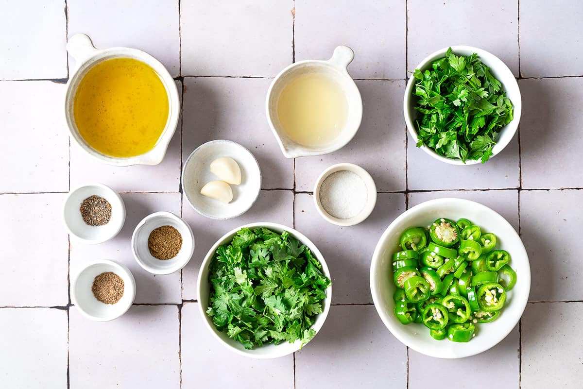 Ingredients for zhoug including sliced jalapeno peppers, garlic, salt, cilantro, parsley, cumin, coriander, cardamom, olive oil and lemon juice.