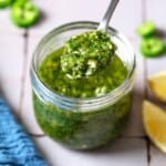 A spoonful of zhoug being held over a jar of the zhoug, with a kitchen towel, sliced jalapenos and lemon wedges in the background.