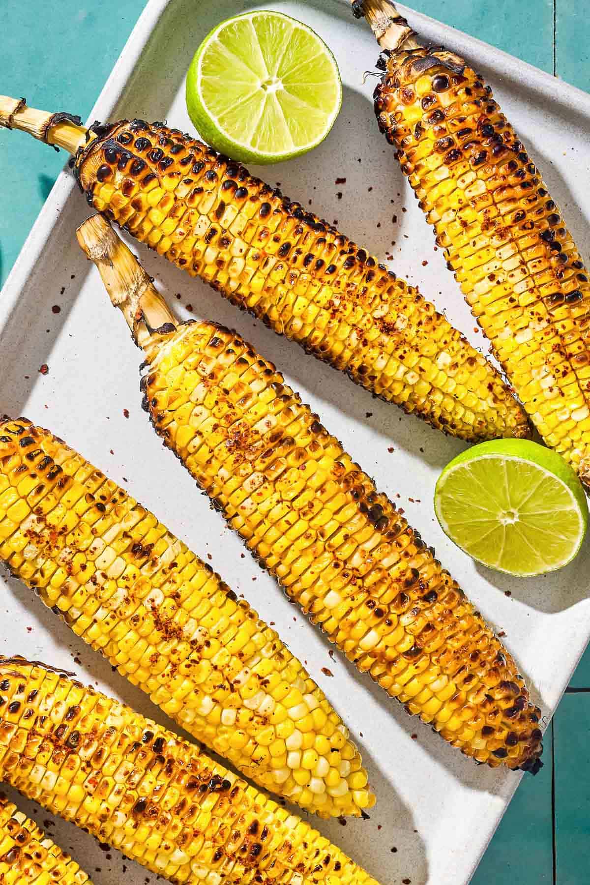 An overhead photo of 6 grilled corncobs on a serving platter with 2 lime halves.