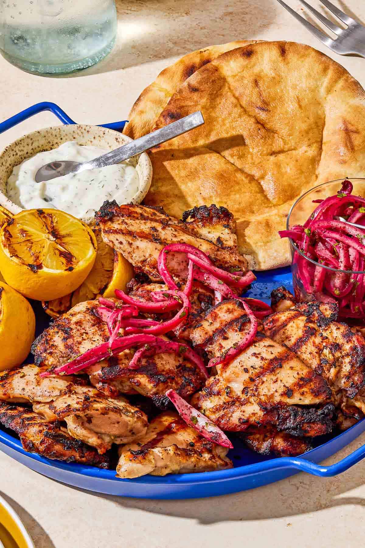 Grilled sumac chicken thighs on a serving platter with pickled red onions, grilled lemon halves, tzatziki sauce, and pita bread.