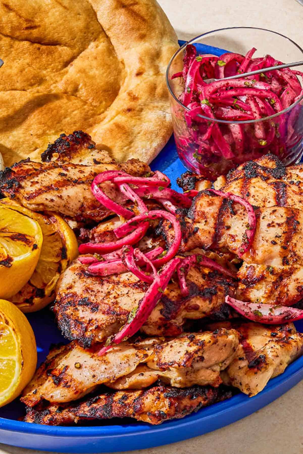 A close up of grilled sumac chicken thighs on a serving platter with pickled red onions, grilled lemon halves, tzatziki sauce, and pita bread.