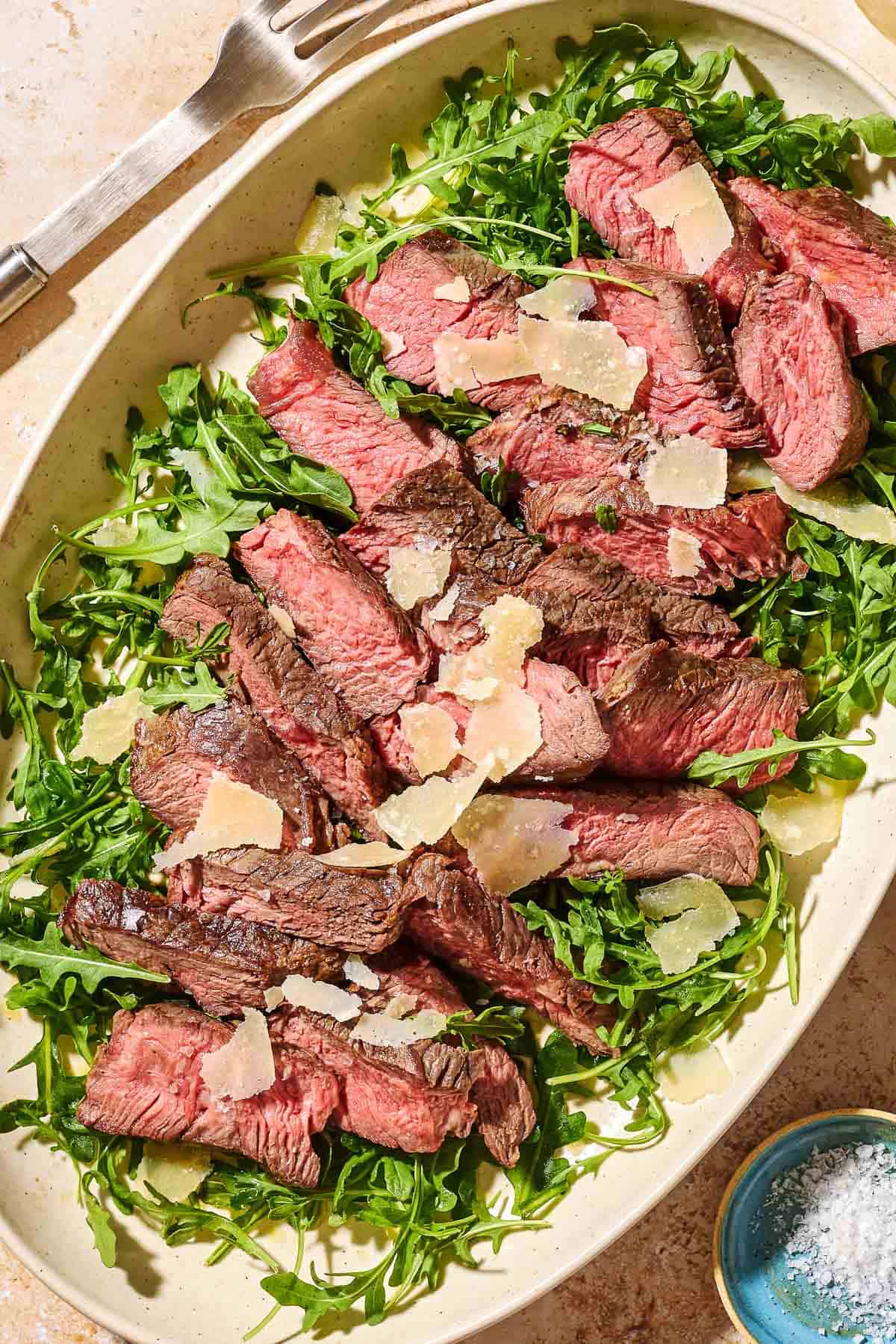 An overhead photo of beef tagliata aka tagliata di manzo topped with shaved parmesan on a bed of arugula on a serving platter. Next to this is a small bowl of salt and a serving fork.
