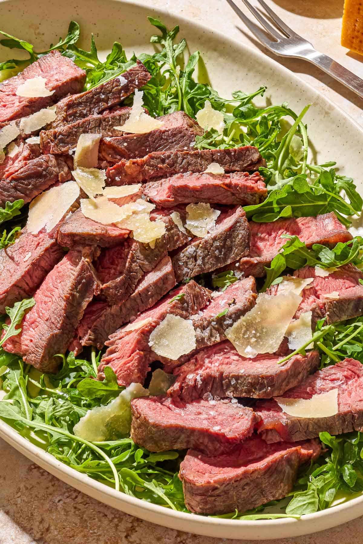 A close up of beef tagliata topped with shaved parmesan on a bed of arugula on a serving platter next to a serving fork.