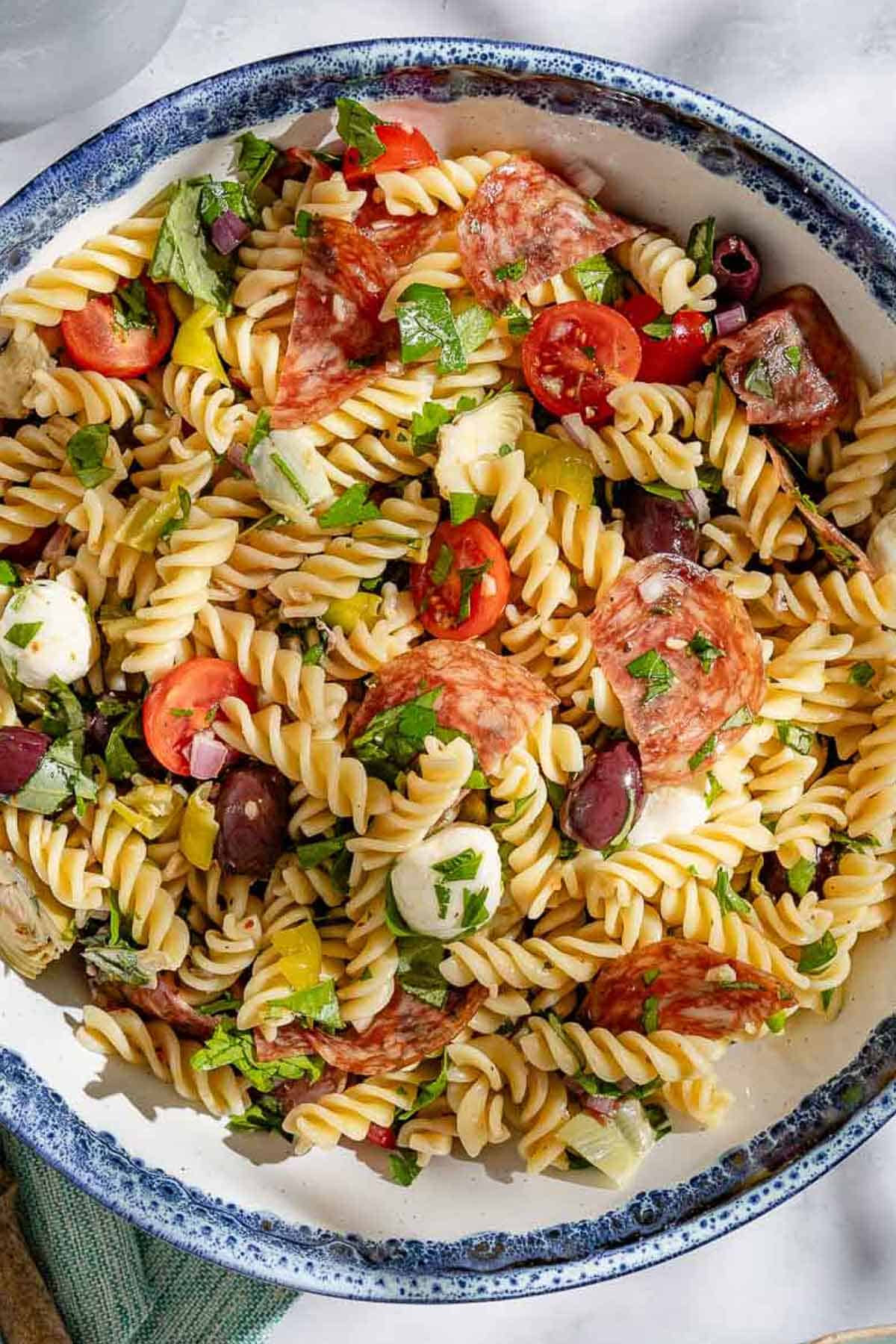 Overhead shot of Italian pasta salad in a serving bowl, showing the mozzarella, tomatoes, pepperoncini, fresh herbs, olives, salami, and artichoke hearts.