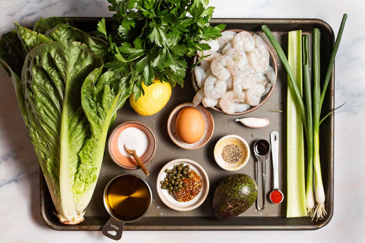 Ingredients for shrimp salad, including shrimp, celery, paprika, worcestershire, parsley, romaine, lemon, egg, salt, pepper, dijon, capers, garlic, olive oil, and avocado.