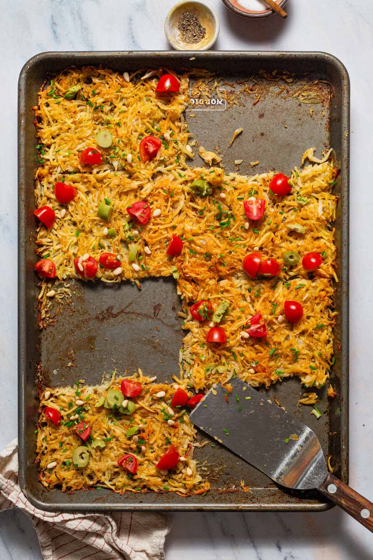 An overhead photo of baked hash browns in a sheet pan with a spatula with 3 pieces missing. Surrounding this is a kitchen towel and bowls of salt and pepper.