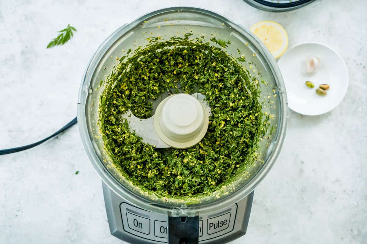 An overhead photo of carrot top pesto in the bowl of a food processor. Next to this is a lemon half and a bowl with two pistachios.