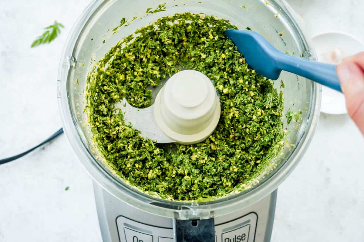 An overhead photo of someone using a spatula to scrap the sides of the carrot top pesto in the bowl of a food processor.