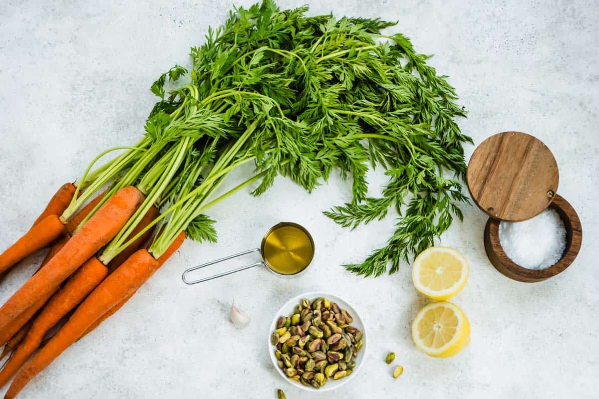 Ingredients for carrot top pesto including carrots with their tops, garlic, pistachios, lemon, olive oil and kosher salt.