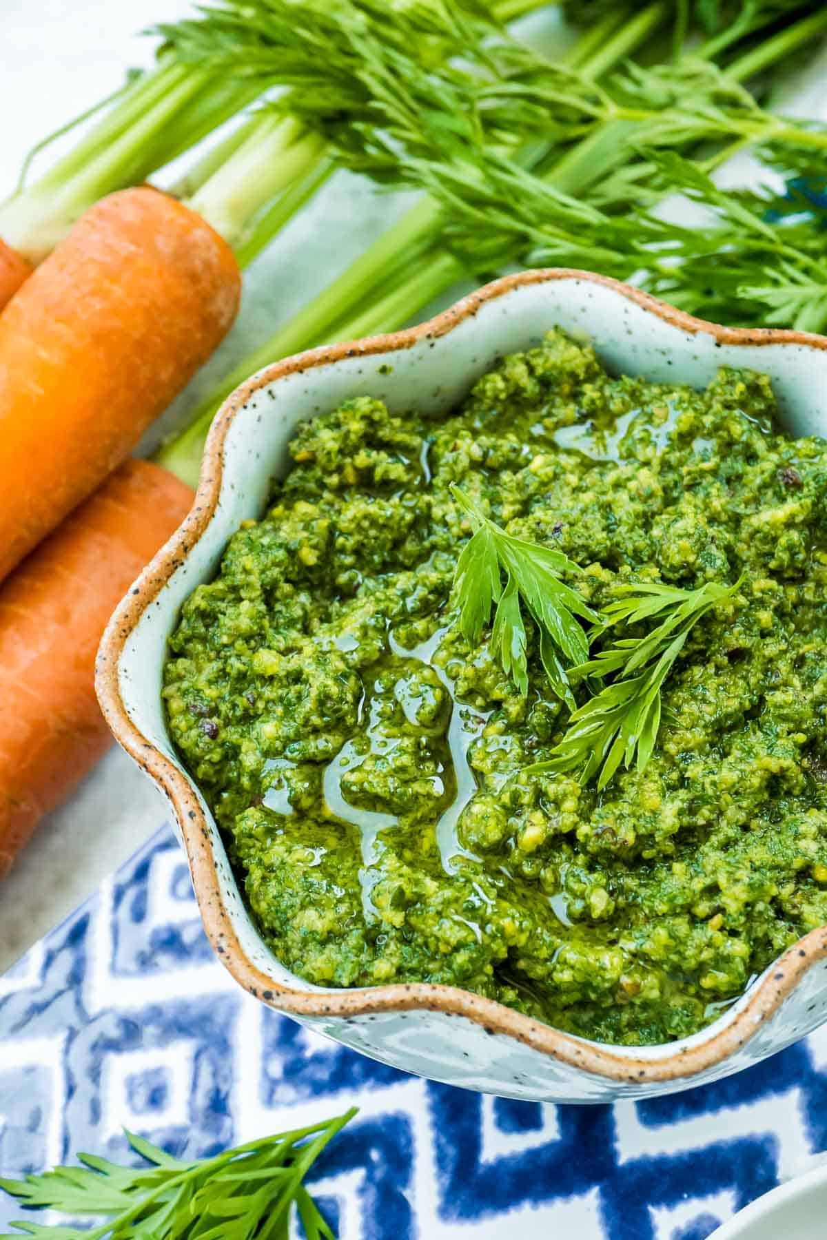 Carrot top pesto in a bowl next to some carrots and bowls of salt and pistachios.