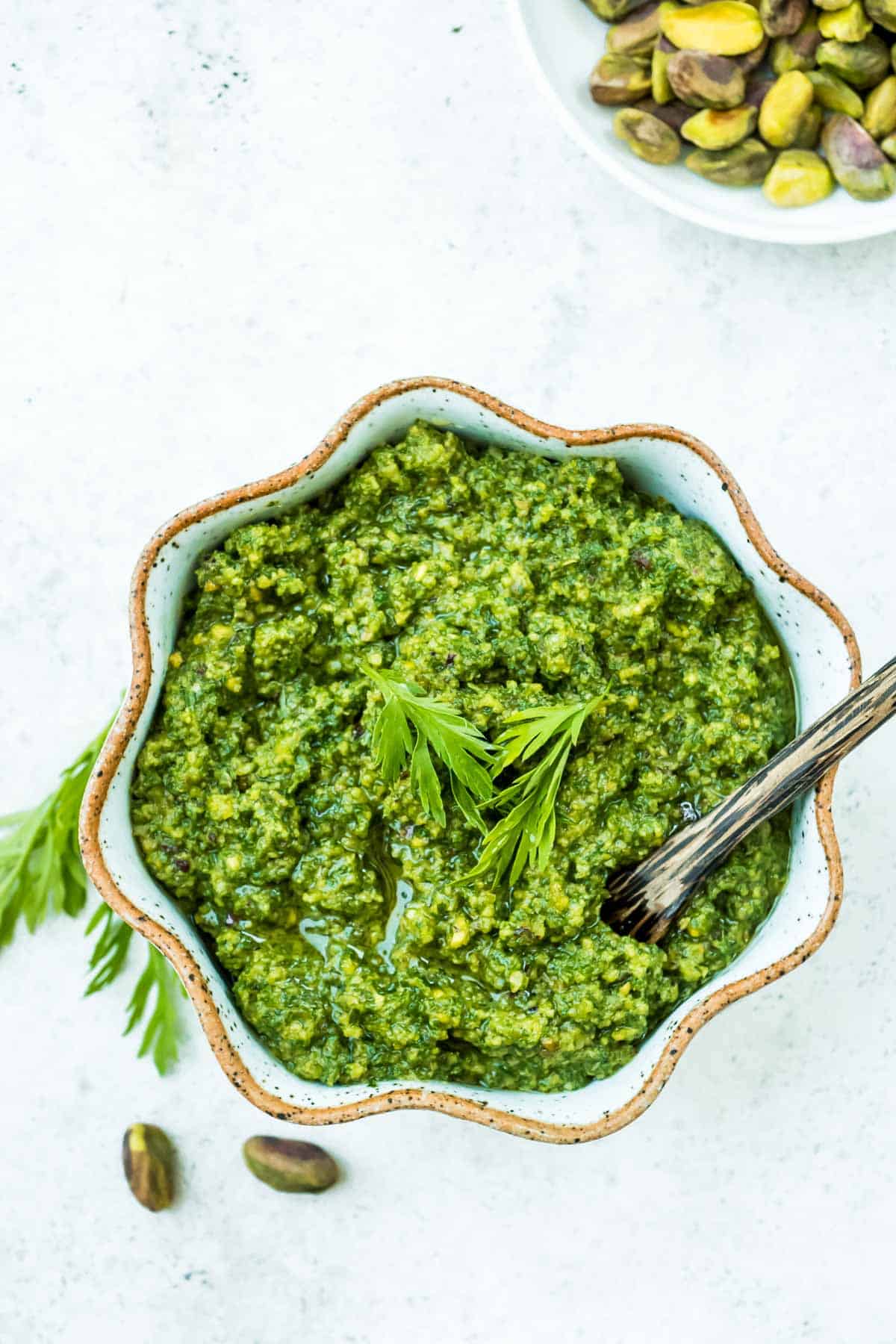 An overhead photo of carrot top pesto in a bowl with a spoon next to a bowl of pistachios.