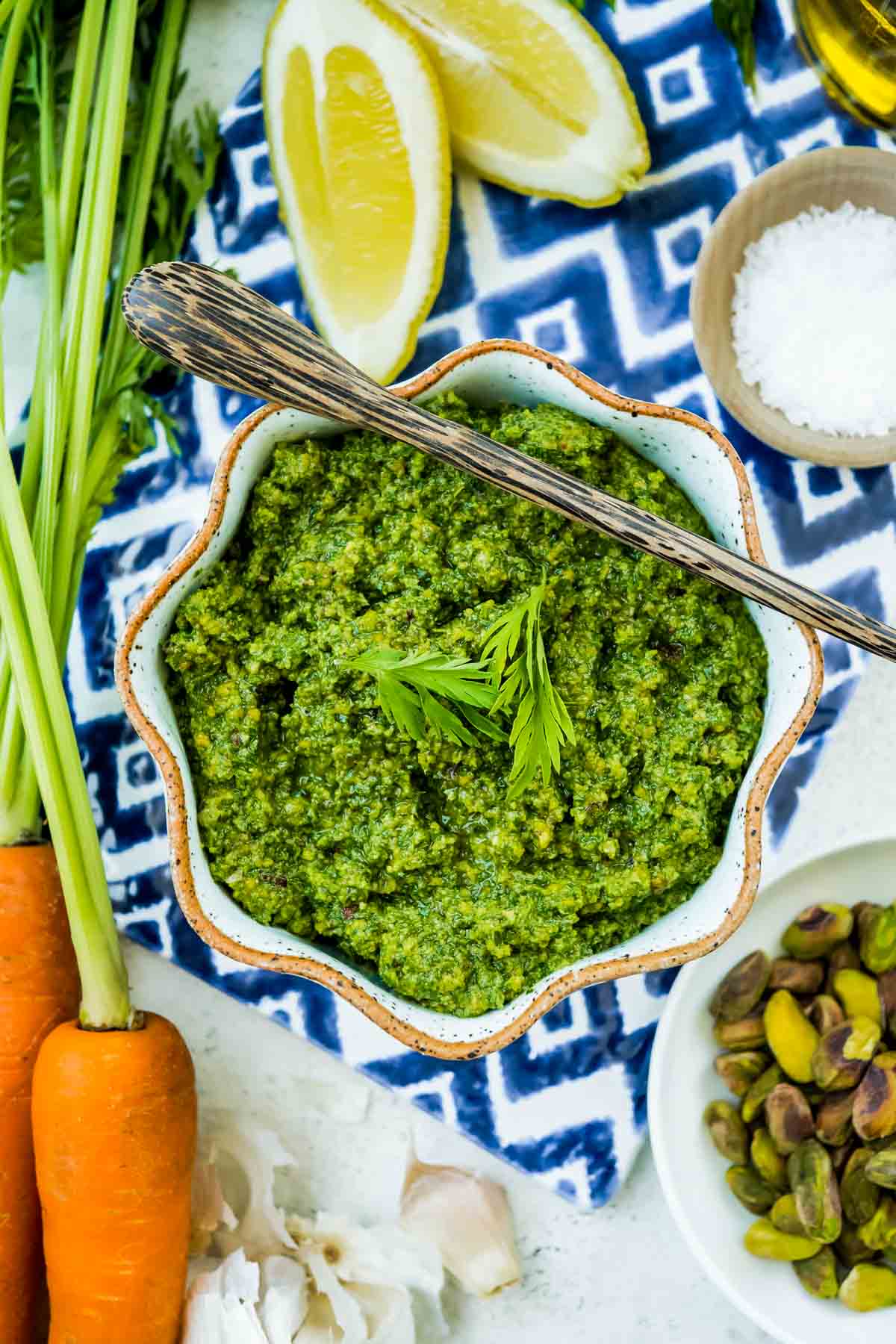 An overhead photo of a bowl of carrot top pesto. Next to this is a bunch of carrots, 2 lemon wedges, a bottle of olive oil, a head of garlic and bowls of salt and pistachios.