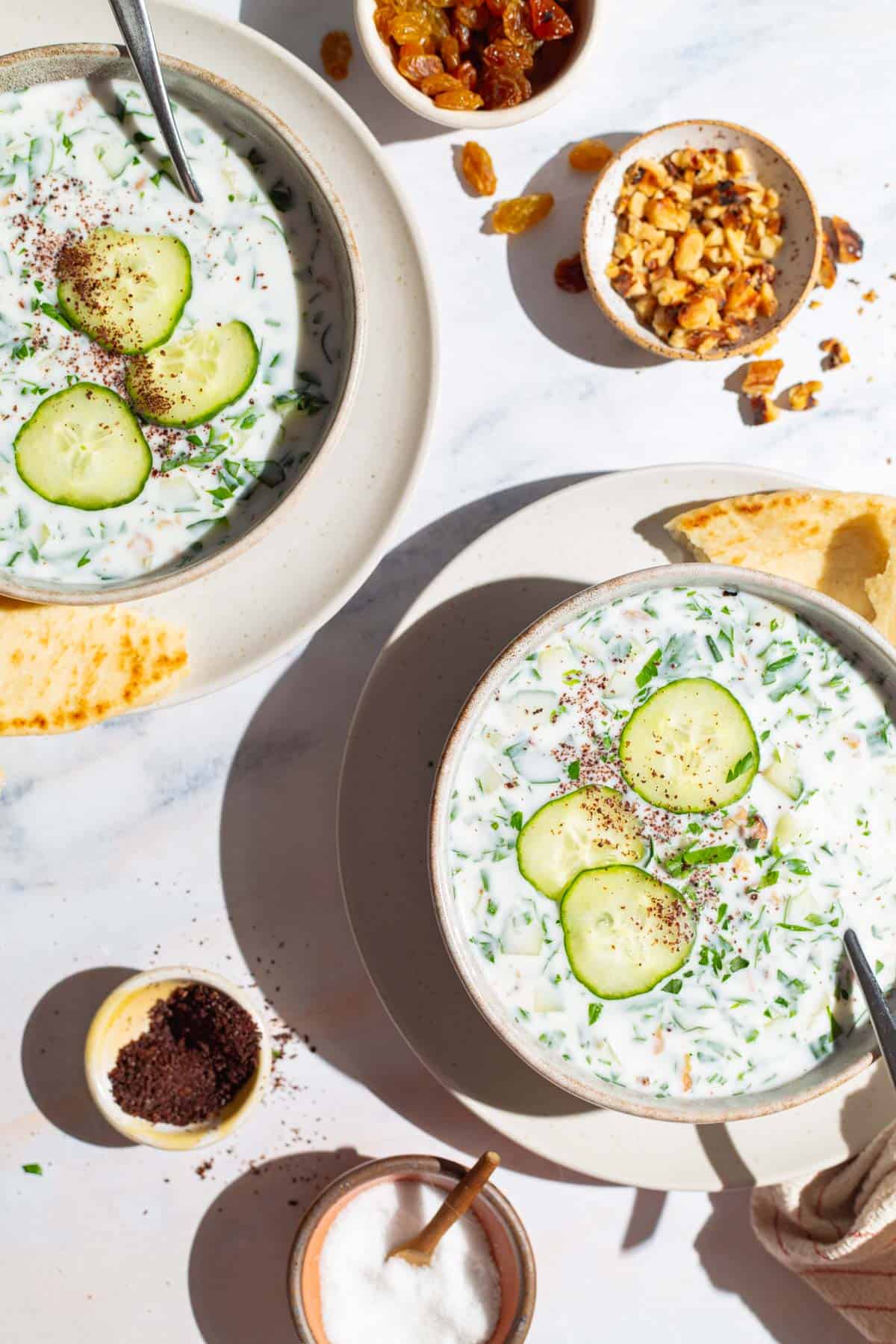 An overhead photo of two bowls of cucumber soup garnished with sumac, parsley and cucumber slices with spoons on plates with slices of pita bread. Next to these are bowls of walnuts, salt, golden raisins and sumac.
