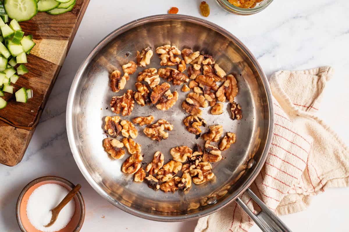 An overhead photo of toasted walnuts in a skillet. Next to this is a kitchen towel, a bowl of salt and a cutting board with chopped cucumber and cucumber slices.